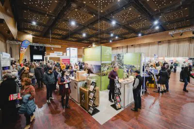 Eine überfüllte Messehalle mit Menschen, die verschiedene Stände und Ausstellungen besuchen. Helle Beleuchtung, hohe Decken und Informationsstände füllen den Raum, während die Besucher mit den Ausstellern interagieren.