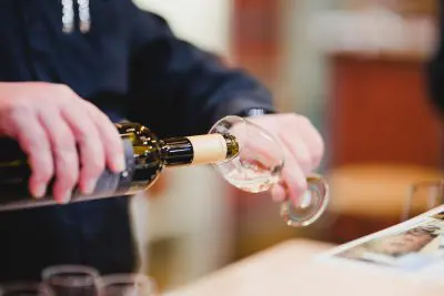 Eine Person in einem dunklen Hemd gießt Weißwein aus einer Flasche in ein klares Weinglas ein. Der Hintergrund ist unscharf und ein bedrucktes Papier ist teilweise auf dem Tisch zu sehen.
