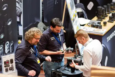 Drei Männer stehen um einen Ausstellungstisch herum und begutachten Kameras und Objektive auf einer Fotomesse, während im Hintergrund verschiedene Kameraausrüstungen und Werbebanner zu sehen sind.
