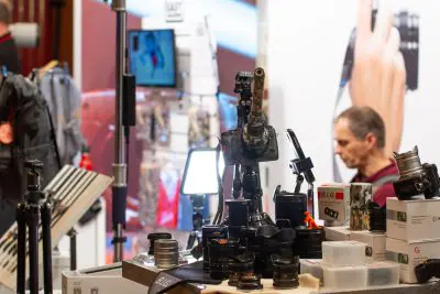 Auf einer Veranstaltung ist ein Ausstellungstisch mit verschiedenen Kameraobjektiven und Zubehör, darunter eine Kamera auf einem Stativ, aufgebaut. Im Hintergrund ist ein Mann mit kurzen Haaren zu sehen, und hinter ihm befinden sich unscharfe Werbeständer.