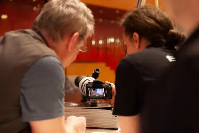Zwei Personen sehen sich eine Digitalkamera mit großem Objektiv genau an und prüfen die Bilder auf dem Bildschirm. Der Hintergrund ist unscharf und weist warme Licht- und Rottöne auf.