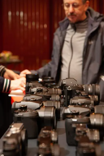 Ein mit verschiedenen Digitalkameras bedeckter Tisch im Vordergrund, im Hintergrund stehen zwei Personen, von denen eine teilweise sichtbar und eine unscharf ist. Die Umgebung scheint ein Innenraum zu sein, möglicherweise auf einer Veranstaltung oder Ausstellung.