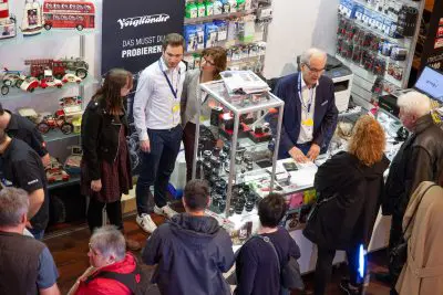 Menschen versammeln sich an einem Messestand um eine Glasvitrine mit Kameras und Objektiven, während die Mitarbeiter ihnen helfen. In den Regalen im Hintergrund stehen Modellautos und Kamerazubehör.