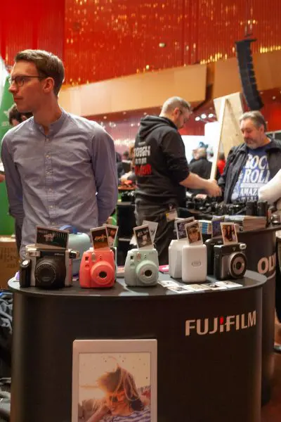 Ein Mann steht neben einem Display mit bunten Fujifilm Instax-Kameras, auf denen Beispielfotos zu sehen sind. Im Hintergrund stöbern Menschen bei einer Veranstaltung in einer Halle mit roter Dekoration.