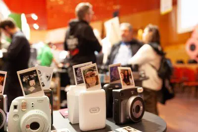 Eine Nahaufnahme von Sofortbildkameras und gedruckten Fotos auf einem Tisch bei einer Veranstaltung in einem Gebäude, mit Menschen, die sich unterhalten und im Hintergrund verschwommen sind. Die Szene ist hell beleuchtet und ungezwungen.