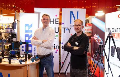 Zwei lächelnde Männer stehen mit verschränkten Armen an einem Messestand, der Fotoausrüstung ausstellt. Bunte Banner und Kameraausrüstung sind zu sehen, mit einer leuchtend roten Wand im Hintergrund.