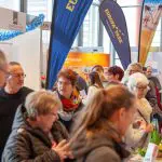 Eine belebte Indoor-Veranstaltung mit Menschen verschiedenen Alters, die an den Ständen herumlaufen und sich unterhalten. Im Hintergrund sind leuchtende Banner und Schilder, darunter auch der Europa-Park, zu sehen. Die Atmosphäre wirkt lebendig und gesellig.