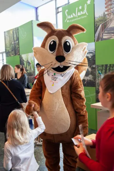 Eine Person in einem braunen Eichhörnchen-Maskottchenkostüm schüttelt einem jungen Mädchen bei einer Indoor-Veranstaltung die Hand. Im Hintergrund sind Menschen und grüne saarländische Werbedisplays zu sehen.