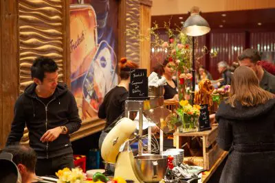 Eine geschäftige Marktszene mit Menschen, die an einer dekorierten Holztheke Essen bestellen und zubereiten, mit Blumen, einem Mixer und Speisekarten auf Kreidetafeln, mit bunten Lichtern und einem alten Plakat im Hintergrund.