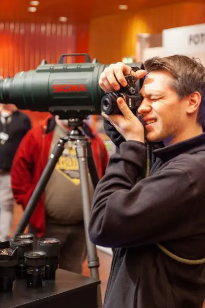 Eine Person macht ein Foto mit einer Kamera, die an einem großen Sigma-Teleobjektiv auf einem Stativ befestigt ist, bei einer Veranstaltung in einer Halle, wobei die Kameraobjektive auf einem Tisch in der Nähe ausgestellt sind.