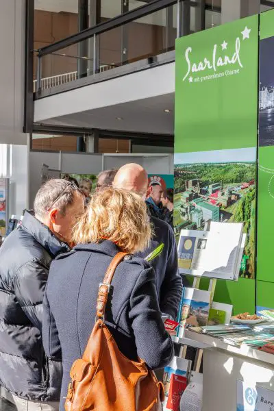 Menschen stehen an einem hell erleuchteten Tourismusstand, der für das Saarland wirbt, mit Broschüren auf dem Tresen und einem grünen Schild mit dem Namen der Region und einem Luftbild im Hintergrund.