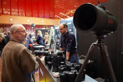 Zwei Männer diskutieren über Kameraobjektive an einem Fotomessestand. Auf einem Tisch sind zahlreiche Kameraobjektive ausgestellt, im Vordergrund ist ein großes Sigma-Teleobjektiv auf einem Stativ montiert. Im Hintergrund sind andere Besucher zu sehen.