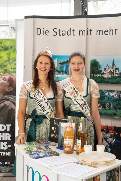 Zwei Frauen in traditionellen bayerischen Kleidern und Schärpen stehen hinter einem Stehtisch mit Getränken, Broschüren und Fotos, vor einem Display, das für eine Stadt wirbt. Beide Frauen lächeln in die Kamera.