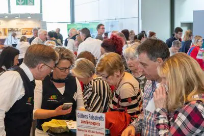 Eine Gruppe von Menschen versammelt sich dicht um einen Stand auf einer Indoor-Veranstaltung oder -Messe, wobei viele die Gegenstände auf einem Tisch begutachten. Ein Schild über Reiseangebote in deutscher Sprache ist zu sehen, und die Atmosphäre wirkt geschäftig und engagiert.