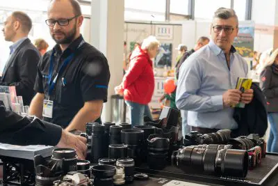 Ein Mann steht hinter einem Ausstellungstisch mit Kameraobjektiven und -ausrüstung bei einer geschäftigen Veranstaltung in einer Halle, während ein anderer Mann mit Brille Broschüren in der Hand hält. Im Hintergrund sind weitere Personen und Stände zu sehen.