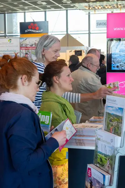 Vier Erwachsene stehen an einem Informationsstand und sehen sich Broschüren und Faltblätter an. Sie scheinen sich für Reise- oder Tourismusmaterial zu interessieren, mit bunten Auslagen und Schildern im Hintergrund.