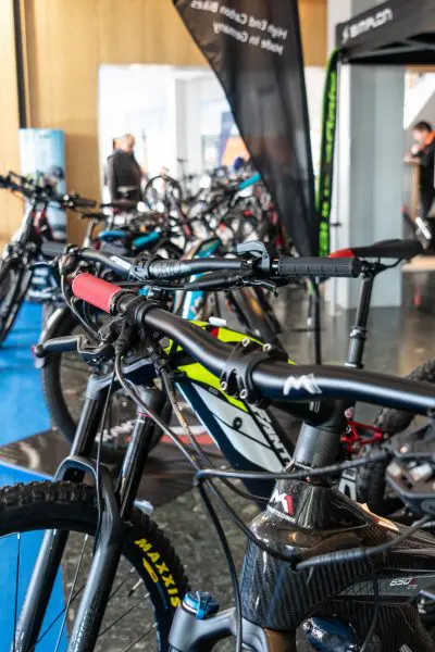 Nahaufnahme des Lenkers eines schwarz-roten Mountainbikes auf einer Indoor-Bike-Messe, mit mehreren anderen Fahrrädern und Menschen im Hintergrund, die unscharf abgebildet sind.