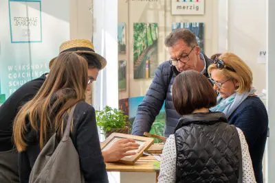 Vier Erwachsene und ein Kind versammeln sich um einen Tisch und betrachten gerahmte Gegenstände und Pflanzen. Sie scheinen an einem Workshop oder einer Diskussion in einem hellen Innenraum mit Informationsplakaten im Hintergrund beteiligt zu sein.
