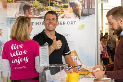 Ein Mann in einem schwarzen Hemd zeigt den Daumen nach oben und lächelt an einem Stand für eine Indoor-Veranstaltung, während eine Frau in einem rosafarbenen "Live Relax Enjoy"-Shirt und ein weiterer Mann mit Papieren in der Nähe stehen. Werbedisplays und Broschüren sind zu sehen.