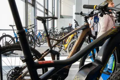 Eine Reihe von Elektrofahrrädern wird in einem hellen, modernen Ausstellungsraum durch große Fenster ausgestellt, während im Hintergrund Menschen die Fahrräder begutachten.