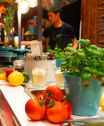 Frische Tomaten und ein Topf mit Basilikum auf einer Küchentheke mit buntem Gemüse, Utensilien und Gläsern; im unscharfen Hintergrund arbeitet eine Person.