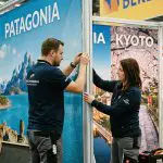 Zwei Personen in Marineuniformen richten einen Ausstellungsstand mit großen Postern der Berge Patagoniens und der Kirschblüten von Kyoto im Hintergrund ein. Sie scheinen eine Tafel für eine Veranstaltung oder Ausstellung zu installieren oder einzustellen.