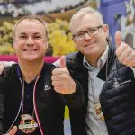 Zwei lächelnde Männer posieren zusammen, beide zeigen den Daumen nach oben. Sie tragen Jacken und große dekorative Halsketten und stehen vor einem bunten, verschwommenen Hintergrund.