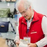 Ein älterer Mann mit grauen Haaren und Brille, der eine rote Weste und ein Namensschild trägt, lächelt, während er eine kleine Schachtel in einem Geschäft hält, in dem hinter ihm Taschen und Accessoires in Regalen ausgestellt sind.