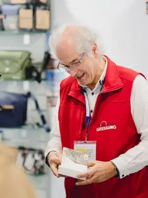 Ein älterer Mann mit grauen Haaren und Brille, der eine rote Weste und ein Namensschild trägt, lächelt, während er eine kleine Schachtel in einem Geschäft hält, in dem hinter ihm Taschen und Accessoires in Regalen ausgestellt sind.