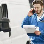 Ein Zusteller in einer blauen Jacke steht draußen neben einem weißen Lieferwagen, lächelt, telefoniert und hält ein Klemmbrett mit Papieren in der Hand.