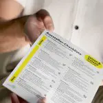 Eine Person in einem weißen Hemd hält eine gedruckte Aussteller-Checkliste mit geordneten Abschnitten und Aufzählungspunkten in deutscher Sprache für die Ausstellungsplanung und -vorbereitung.