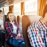 Eine junge Frau mit Kopfhörern und einem karierten Hemd sitzt lächelnd in einem Bus. Andere Fahrgäste sitzen in der Nähe. Das Sonnenlicht strömt durch die Fenster und schafft eine warme Atmosphäre.