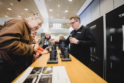 Mehrere Personen untersuchen Kameraobjektive, die auf einem Holztisch ausgestellt sind. Sie werden dabei von einem Vertreter unterstützt, der ein Tamron-Hemd auf einer Fotoveranstaltung oder Messe trägt.