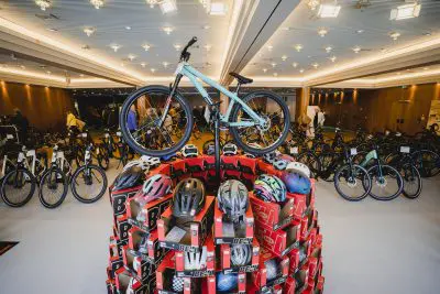Ein blaugrünes Fahrrad steht auf einem kuppelförmigen Stapel roter Kisten mit verschiedenen Helmen in einem großen, gut beleuchteten Ausstellungsraum, der mit weiteren Fahrrädern und Menschen im Hintergrund gefüllt ist.