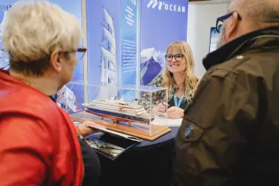 Eine lächelnde Frau sitzt hinter einem Tisch, auf dem ein Schiffsmodell ausgestellt ist, und unterhält sich mit zwei Personen an einem Veranstaltungsstand mit Postern zum Thema Meer im Hintergrund.