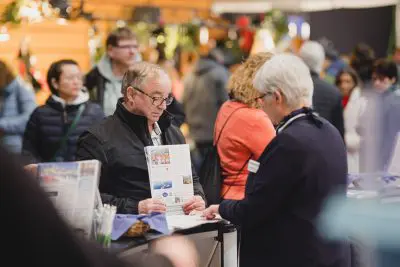 Ein Mann, der eine Broschüre in der Hand hält, spricht mit einer Frau an einem überdachten Informationsstand, während sich im Hintergrund weitere Personen versammeln und unterhalten. Die Umgebung wirkt belebt und gut beleuchtet.
