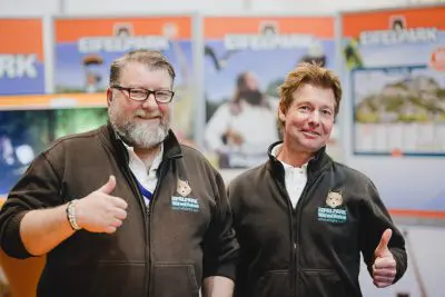 Zwei Männer in braunen Eifelpark-Jacken lächeln und zeigen den Daumen nach oben. Sie stehen in einem Innenraum mit Eifelpark-Werbeplakaten und Aufstellern im Hintergrund.