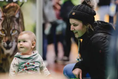 Eine Frau mit einer schwarzen Wintermütze lächelt ein Baby an, das einen hellen Kapuzenpulli trägt. Sie befinden sich in einem Innenraum, vor einem unscharfen Hintergrund mit Menschen und einem Poster, das einen Teil des Gesichts einer Wildkatze zeigt.