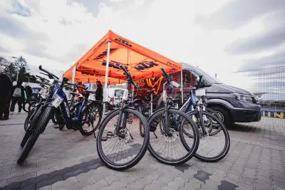 Unter einem orangefarbenen Außenzelt mit KTM-Schriftzug sind mehrere Fahrräder ausgestellt. Die Menschen sind versammelt, und ein großer grauer Lieferwagen ist in der Nähe auf einem gepflasterten Platz unter einem bewölkten Himmel geparkt.