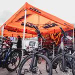 Unter einem leuchtend orangefarbenen KTM-Zelt werden bei einer Outdoor-Veranstaltung mehrere Fahrräder ausgestellt, während im Hintergrund weitere Fahrräder und Ausrüstung zu sehen sind.