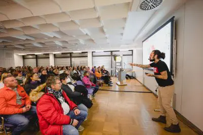 Ein Mann mit Bart und Pferdeschwanz hält eine lebhafte Präsentation vor einem sitzenden Publikum in einem modernen Konferenzraum und deutet mit einer Geste auf einen großen Bildschirm, auf dem bunte Bilder zu sehen sind.