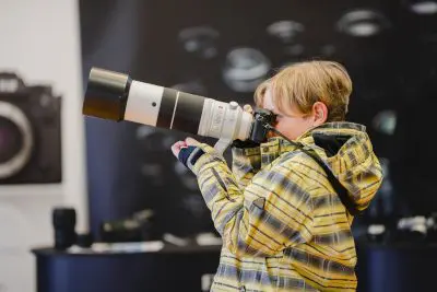 Ein Kind in einer gelben karierten Jacke hält eine große Kamera mit einem langen Teleobjektiv in der Hand und schaut durch sie hindurch, wobei es in einem Innenraum mit unscharfer Kameraausrüstung im Hintergrund nach vorne schaut.