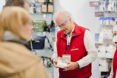 Ein älterer Mann mit weißen Haaren, der eine Brille und eine rote Weste trägt, zeigt den Kunden in einem Geschäft ein Produkt. Im Hintergrund sind Regale mit Taschen und verschiedenen Artikeln zu sehen.