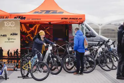 Zwei Personen begutachten Fahrräder vor einem orangefarbenen KTM-Zelt bei einer Outdoor-Veranstaltung, umgeben von mehreren Fahrrädern und Bannern, und einem in der Nähe geparkten Lieferwagen.