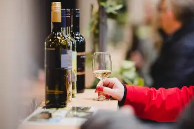 Eine Person in einer roten Jacke hält ein Glas Weißwein an einem hölzernen Tresen mit mehreren Weinflaschen; andere Personen sind unscharf im Hintergrund zu sehen.