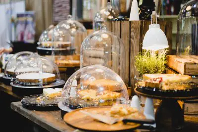 In einer warm beleuchteten Bäckerei oder einem Café werden verschiedene Torten und Kuchen auf Holztabletts und Ständern mit Glaskuppeln ausgestellt. Eine kleine Lampe und eine Rosmarinpflanze tragen zur gemütlichen Atmosphäre bei.