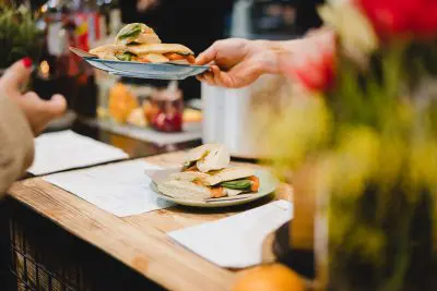 Eine Person reicht einer anderen Person über einem Holztresen einen Teller mit einem mit Spinat, Tomaten und Käse belegten Sandwich, während im Vordergrund ein weiteres ähnliches Sandwich auf einem Teller liegt.