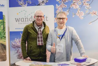 Zwei lächelnde ältere Männer stehen an einem Informationsstand mit Broschüren und Stiften. Der eine Mann trägt eine Brille, einen grünen Pullover und einen Schal, der andere, ebenfalls mit Brille, zeigt den Daumen nach oben. Hinter ihnen befinden sich eine Blumenkulisse und ein Wanderschild.