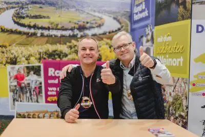Zwei Männer sitzen nebeneinander an einem Tisch, lächeln und zeigen die Daumen nach oben. Sie tragen Medaillen um ihren Hals. Hinter ihnen ist eine bunte Kulisse mit malerischen Fluss- und Landschaftsfotos sowie Tourismusplakaten zu sehen.