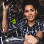 Eine junge Frau mit lockigem Haar und einem karierten Hemd lächelt, während sie den Lenker eines Fahrrads in einem Laden voller anderer Fahrräder hält.